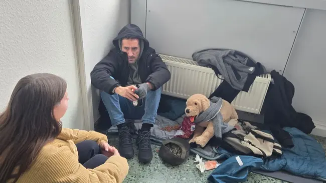 Eine Frau spricht mit einem Mann, der auf dem Boden sitzt und scheinbar in einer schwierigen Lebenssituation ist. Neben ihm liegt ein Teddybär und zerknüllte Kleidung. Die Atmosphäre wirkt ernst und bedürftig.
