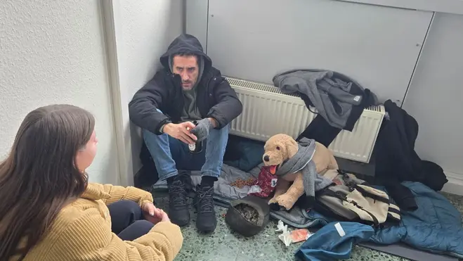 Eine Frau spricht mit einem Mann, der auf dem Boden sitzt und scheinbar in einer schwierigen Lebenssituation ist. Neben ihm liegt ein Teddybär und zerknüllte Kleidung. Die Atmosphäre wirkt ernst und bedürftig.