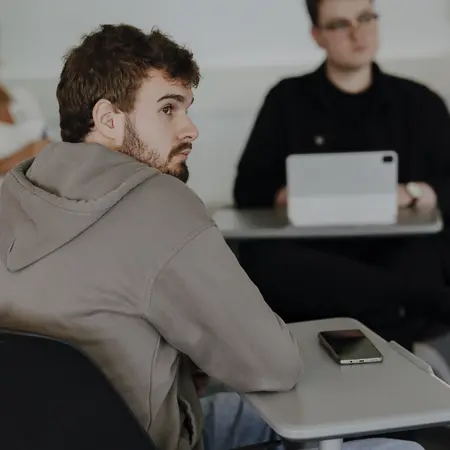Man sieht im Vordergrund einen jungen Mann, der an einem Schreibtisch sitzt und aufmerksam zuhört. Im Hintergrund sitzen zwei andere Personen, die in die gleiche Richtung schauen und zuhören.