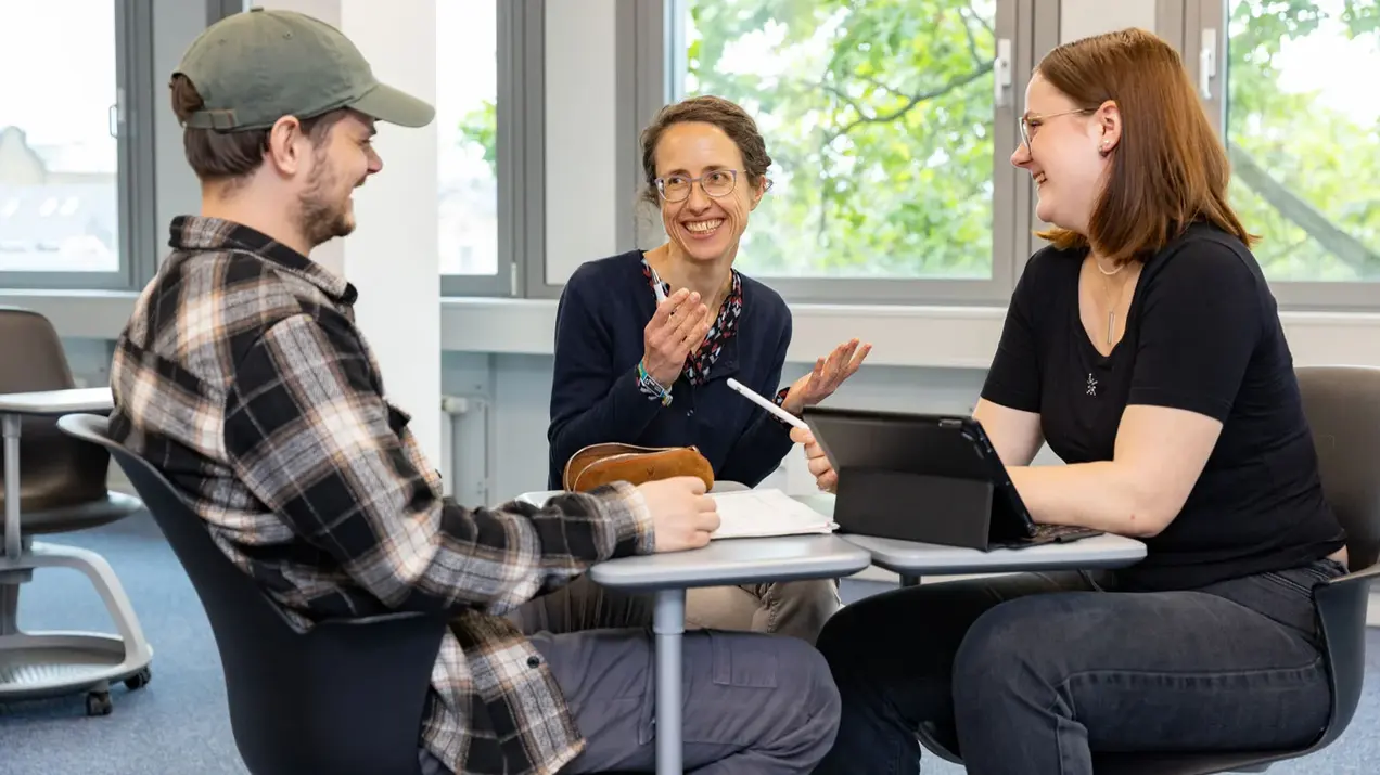 Zwei Studierende sitzen sich an zwei schmalen Tischen gegenüber und simulieren ein Bewerbungsgespräch, zwischen Ihnen gibt eine Beraterin Tipps