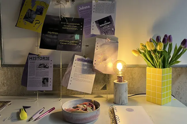 Ein kreativ gestalteter Schreibtisch mit einem Drahtdisplay, auf dem verschiedene Dokumente und Artikel hängen. Neben dem Display steht eine gelbe Vase mit lila Tulpen und eine Lampe leuchtet warm. Im Vordergrund sind ein Notizbuch und eine Schale mit bunten Gummibärchen sichtbar.