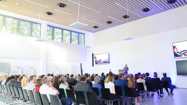 Eine große Gruppe von Menschen sitzt in einem modernen Raum und hört Prof. Dr. Barbara Seegebarth zu, die auf einer Bühne steht. 