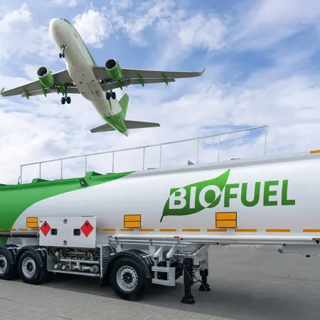 Ein grüner Tankwagen mit der Aufschrift „BIOFUEL“ steht auf der Straße, während ein Flugzeug über ihm landet. Der Himmel ist klar, mit einigen Wolken, und im Hintergrund ist eine grüne Wiese zu sehen.
