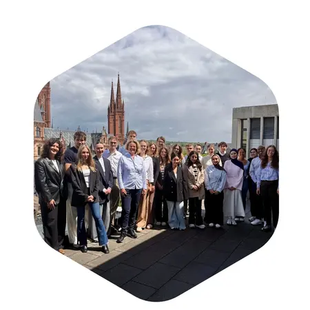 Eine Gruppe von jungen Erwachsenen steht auf einer Dachterrasse mit einer Stadtansicht im Hintergrund. Sie tragen unterschiedliche, formelle Kleidung und lächeln freundlich in die Kamera. Die Kulisse zeigt markante Gebäude und einen bewölkten Himmel.