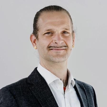 Ein Mann mit langem, umgekämmtem Haar und einem Schnurrbart lächelt in die Kamera. Er trägt ein dunkles Jackett über einem weißen Hemd. Am Ohr hat er goldene Ohrstecker. Der Hintergrund ist hell und neutral.