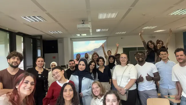 Eine Gruppe von etwa 30 Menschen steht in einem Raum und lächelt. Einige Personen heben die Hände. Sie sind vielfältig gekleidet und scheinen Freude zu haben. Im Hintergrund ist ein Projektor mit einer Präsentation zu sehen.