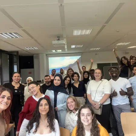 Eine Gruppe von etwa 30 Menschen steht in einem Raum und lächelt. Einige Personen heben die Hände. Sie sind vielfältig gekleidet und scheinen Freude zu haben. Im Hintergrund ist ein Projektor mit einer Präsentation zu sehen.