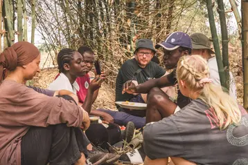 A group of seven people are sitting outdoors under a bamboo grove. They are talking to each other and sharing a meal. The atmosphere is relaxed and convivial, with all participants engaged in lively conversation. They are HSRM students on an excursion and their hosts in Ghana.