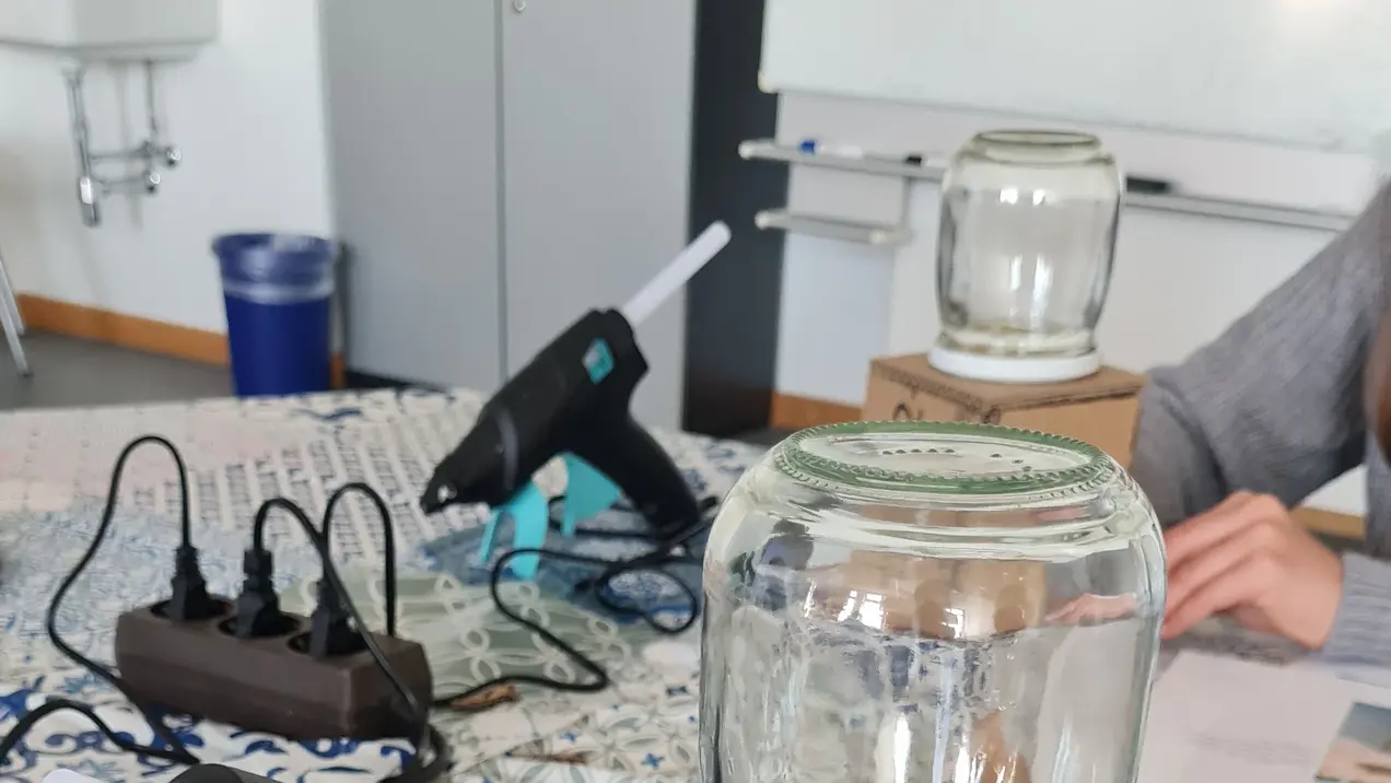Auf dem Tisch stehen mehrere Gläser auf Kartonständern. Eines der Gläser ist transparent und wird mit einem Deckel versehen. Neben dem Glas befindet sich eine Heißkleberpistole und einige Bastelmaterialien. Der Tisch hat ein gemustertes Tischtuch.