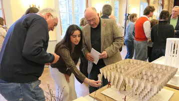 Drei Personen stehen um ein Architekturmodell. Eine Studentin zeigt auf das Modell und erklärt ihren Entwurf, während zwei Männer aufmerksam zuschauen.
