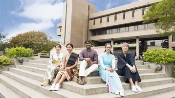 Five people sit on steps in front of a modern building, wearing traditional and modern clothing. Trees and flowering plants are visible; sky partly cloudy.