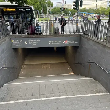 Ein Zugang zur Unterführung am Wiesbadener Hauptbahnhof mit Treppen, die hinunterführen. Oben befinden sich Schilder für Bussteige A und B. Im Hintergrund warten Menschen auf den Bus.