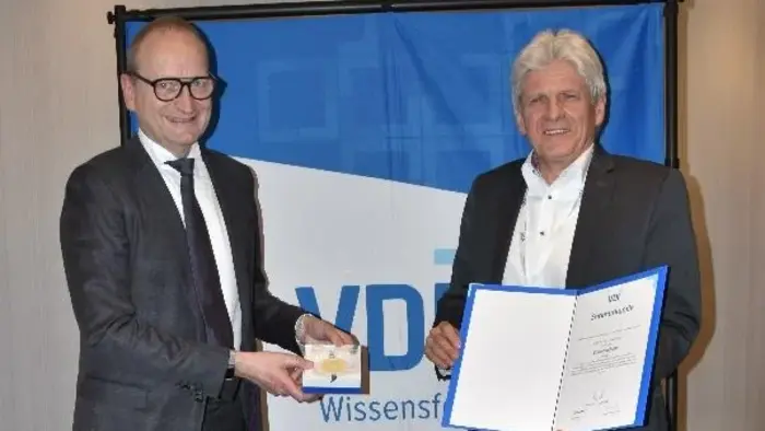 Zwei Männer stehen nebeneinander vor einem blauen Hintergrund mit dem VDI-Logo. Der linke Mann überreicht dem rechten eine Auszeichnung und ein Zertifikat. Beide sind formell gekleidet und lächeln.