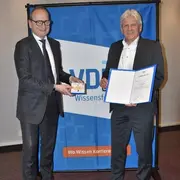 Zwei Männer stehen nebeneinander vor einem blauen Hintergrund mit dem VDI-Logo. Der linke Mann überreicht dem rechten eine Auszeichnung und ein Zertifikat. Beide sind formell gekleidet und lächeln.