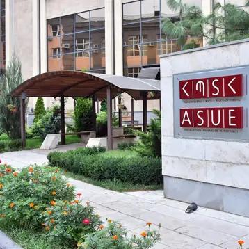 Das Bild zeigt das Schild der Armenischen Staatsuniversität für Wirtschaft, das den Namen der Institution in mehreren Sprachen präsentiert. Im Hintergrund sind Pflanzen und architektonische Elemente des Campus sichtbar, die einen freundlichen Außenbereich deutlicher machen.