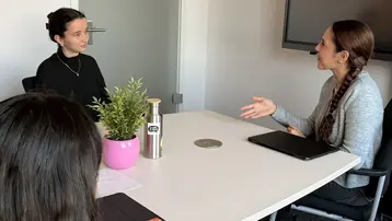 Zwei Frauen führen ein Gespräch an einem Tisch in einem modernen Büroraum. Eine Frau sitzt und hört aufmerksam zu, während die andere aktiv spricht. Auf dem Tisch steht eine Pflanze und eine Wasserflasche.