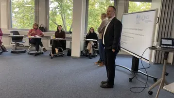 Eine Gruppe von Personen sitzt in einem modernen Klassenraum. Im Vordergrund stehen zwei Männer, einer in Anzug, der andere in lässiger Kleidung. Im Hintergrund ist ein Bildschirm mit Schrift, die ein Thema zur interaktiven Kommunikation präsentiert. Die Fenster zeigen grüne Bäume.