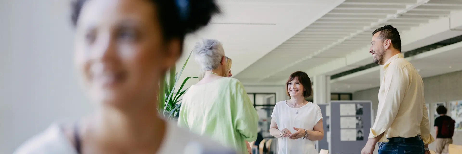 Eine Gruppe von vier Mitarbeitende der Hochschule RheinMain steht in einem hellen Raum und unterhält sich freundlich. Im Vordergrund hält eine Studierende ein Smartphone in der Hand. Die Atmosphäre wirkt entspannt und kommunikativ.