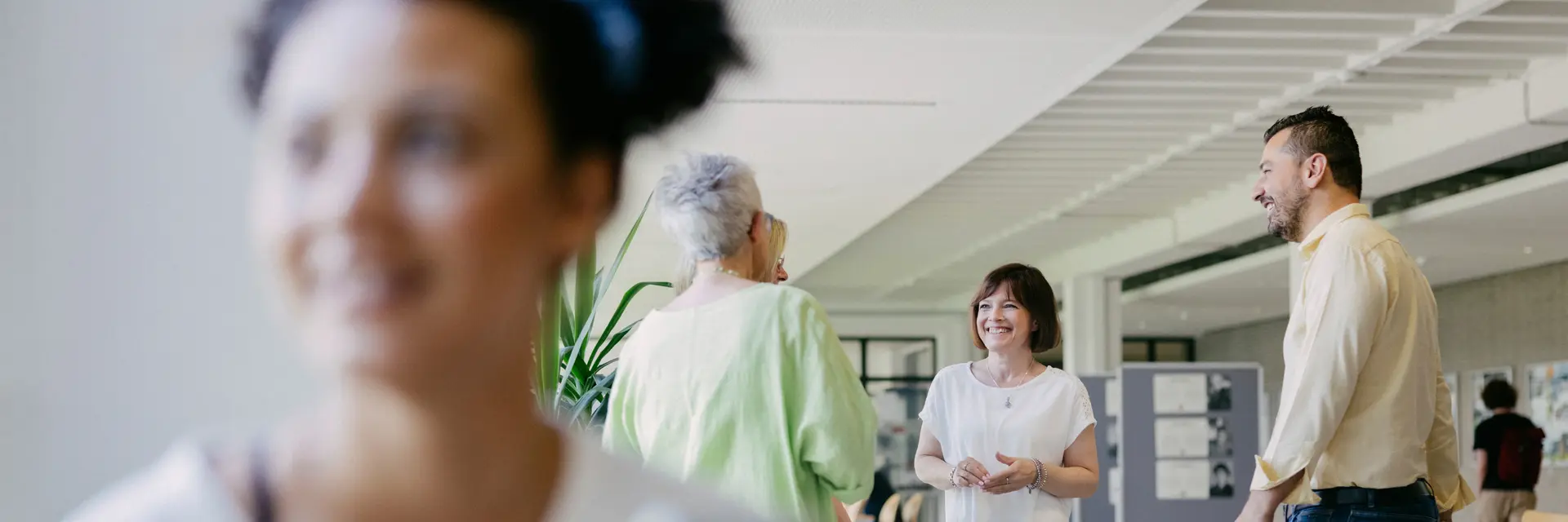 Eine Gruppe von vier Mitarbeitende der Hochschule RheinMain steht in einem hellen Raum und unterhält sich freundlich. Im Vordergrund hält eine Studierende ein Smartphone in der Hand. Die Atmosphäre wirkt entspannt und kommunikativ.