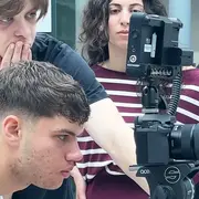 Eine Gruppe von drei Personen beobachtet konzentriert einen Kameramonitor. Ein junger Mann sitzt vor der Kamera, während zwei weitere Personen, eine Frau und ein Mann, hinter ihm stehen und den Monitor betrachten, um das Bild zu überprüfen.