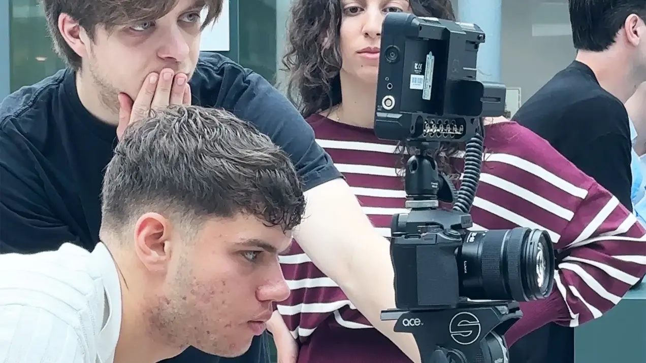 Eine Gruppe von drei Personen beobachtet konzentriert einen Kameramonitor. Ein junger Mann sitzt vor der Kamera, während zwei weitere Personen, eine Frau und ein Mann, hinter ihm stehen und den Monitor betrachten, um das Bild zu überprüfen.
