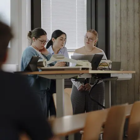 Man sieht drei Studentinnen, die einen gemeinsamen Vortrag halten und dafür vorne an einem Pult stehen und einen Studierenden, der zuhört.