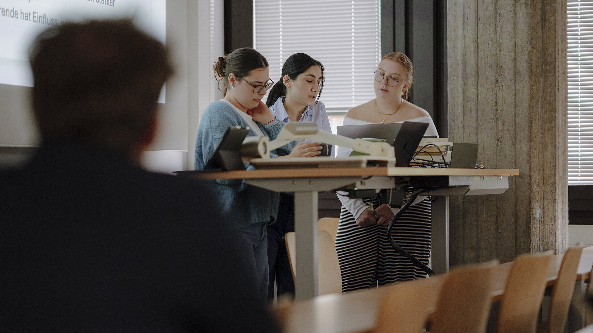 Vortrag KSR Man sieht drei Studentinnen, die einen gemeinsamen Vortrag halten und dafür vorne an einem Pult stehen und einen Studierenden, der zuhört.