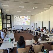 Vortrag/Präsentation vor einer Gruppe Menschen, die Im Kreis an Tischen verteilt sitzt