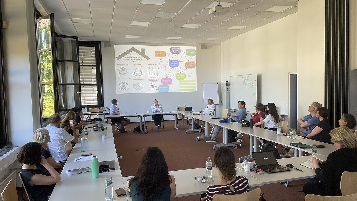Vortrag/Präsentation vor einer Gruppe Menschen, die Im Kreis an Tischen verteilt sitzt