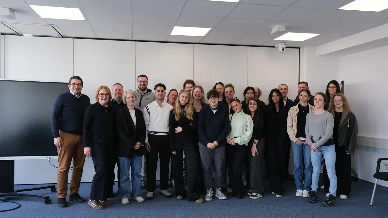 Eine Gruppe von etwa 25 Personen steht in einem Raum und lächelt in die Kamera. Sie posieren in lockerer Kleidung, vor einem großen Bildschirm. Der Raum hat helle Wände und eine professionelle Atmosphäre.