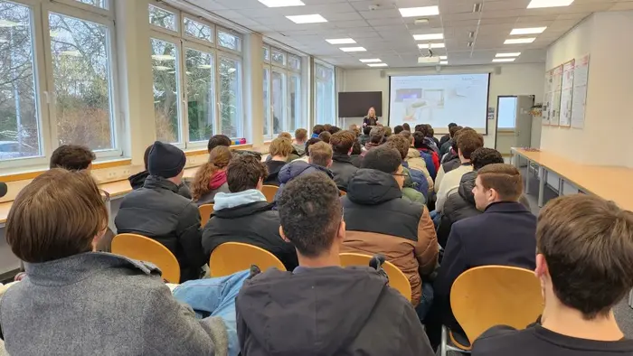 Eine Gruppe von Schülerinnen und Schülern sitzt in einem Raum mit Tischen und Stühlen. Sie sehen auf eine Präsentation an der Wand, während eine Referentin vor ihnen spricht. Große Fenster lassen Tageslicht herein.