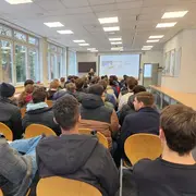 Eine Gruppe von Schülerinnen und Schülern sitzt in einem Raum mit Tischen und Stühlen. Sie sehen auf eine Präsentation an der Wand, während eine Referentin vor ihnen spricht. Große Fenster lassen Tageslicht herein.
