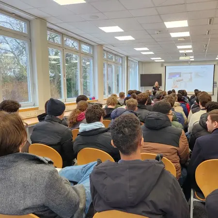 Eine Gruppe von Schülerinnen und Schülern sitzt in einem Raum mit Tischen und Stühlen. Sie sehen auf eine Präsentation an der Wand, während eine Referentin vor ihnen spricht. Große Fenster lassen Tageslicht herein.