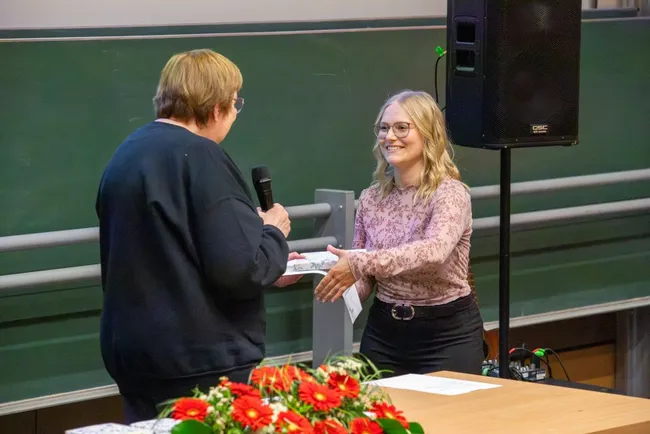 Zwei Frauen stehen sich in einem Raum gegenüber. Eine Frau überreicht der anderen ein Geschenk oder eine Auszeichnung. Auf dem Tisch vor ihnen sind Blumenarrangements zu sehen. Der Raum scheint für eine Veranstaltung oder Zeremonie vorbereitet zu sein.