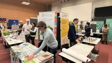 Eine Veranstaltung mit mehreren Ständen, an denen Menschen Informationstafeln und Materialien betrachten. Eine Frau steht an einem Tisch und sortiert Materialien, während mehrere Personen in der Nähe mit Informationsständen beschäftigt sind. Die Umgebung ist freundlich und einladend.