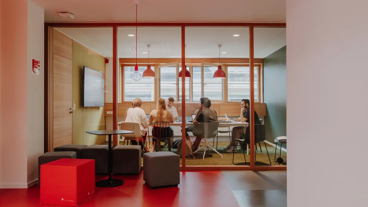 Eine Gruppe von fünf Personen sitzt um einen Tisch in einem Besprechungsraum mit großen Fenstern. Die Atmosphäre ist freundlich, mit modernen Möbeln und warmem Licht. Ein Bildschirm ist an der Wand sichtbar. Die Menschen scheinen in eine Diskussion vertieft zu sein.