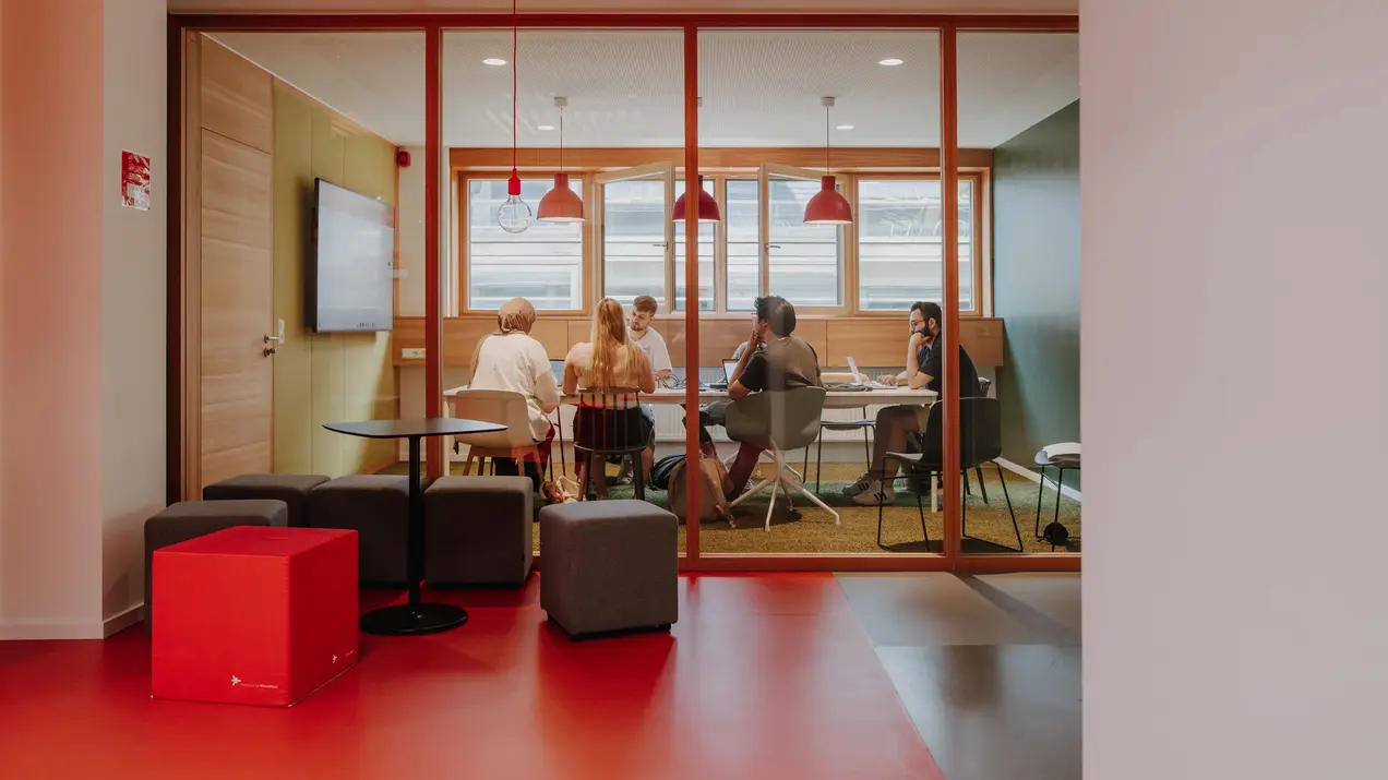 Eine Gruppe junger Menschen sitzt um einen Tisch in einem Besprechungsraum mit großen Fenstern und arbeitet miteinander. Die Atmosphäre ist freundlich, mit modernen Möbeln und warmem Licht. An der Wand hängt ein großer Bildschirm.