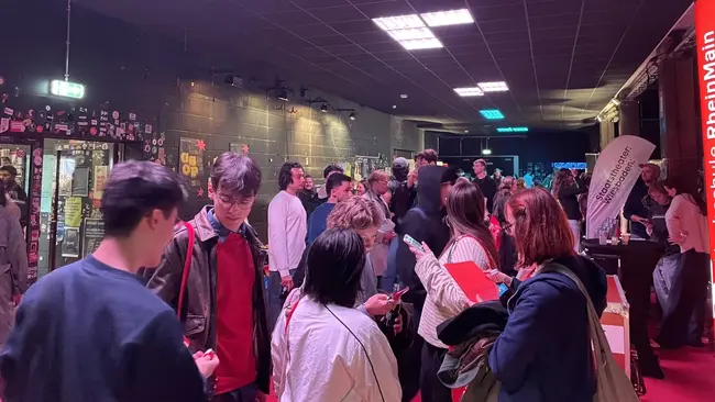 Eine Menschenmenge steht in einem Innenraum, der gedämpft beleuchtet ist. Einige Personen unterhalten sich, während andere etwas in der Hand halten. Im Hintergrund sind Wände mit Aufklebern und Werbematerial sichtbar. Ein Stehtisch mit einem Banner ist ebenfalls im Bild.