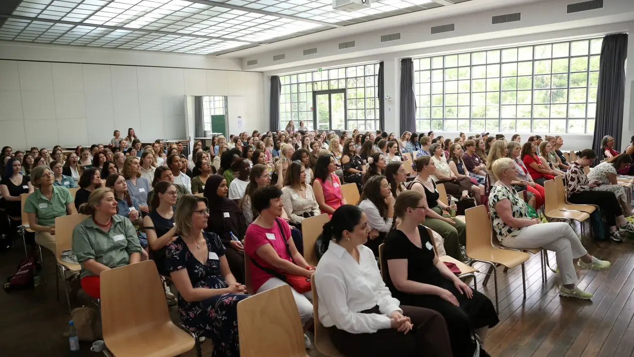 Eine große Gruppe von Menschen sitzt in einem hellen Raum mit großen Fenstern. Die Menge besteht überwiegend aus Frauen, die aufmerksam in Richtung einer nicht sichtbaren Bühne blicken. Einige haben Namensschilder auf ihren Kleidungsstücken.