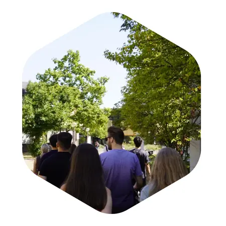 Eine Gruppe von Menschen geht durch eine grüne, baumbestandene Umgebung. Im Vordergrund sind die Rücken der Personen zu sehen, während sie einen schmalen Weg entlang schreiten. Der Himmel ist blau und die Pflanzen wirken frisch und lebendig.