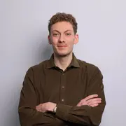 Ein Mann mit lockigem, kurz geschnittenem Haar steht vor einem hellen Hintergrund. Er trägt ein dunkelgrünes Hemd und hat die Arme vor der Brust verschränkt. Er lächelt freundlich und wirkt selbstbewusst.