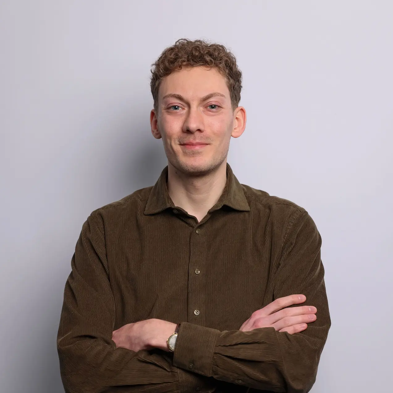 Ein Mann mit lockigem, kurz geschnittenem Haar steht vor einem hellen Hintergrund. Er trägt ein dunkelgrünes Hemd und hat die Arme vor der Brust verschränkt. Er lächelt freundlich und wirkt selbstbewusst.