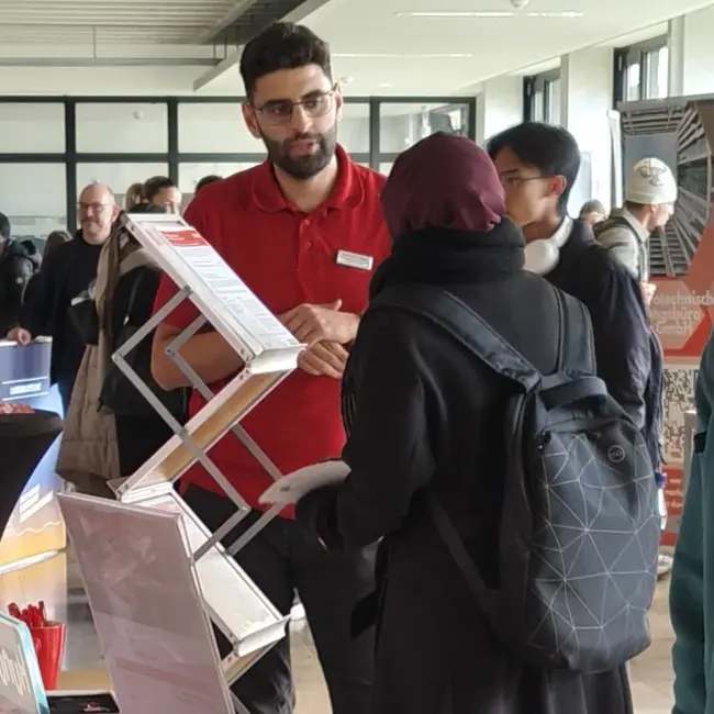 Ein Mann in einem roten Poloshirt erklärt einer Frau mit Kopftuch etwas an einem Informationsstand. Auf dem Tisch stehen verschiedene Broschüren und Informationsmaterialien. Im Hintergrund sind weitere Personen zu sehen, die sich um den Stand versammeln.