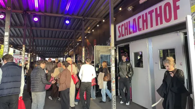 Ein belebter Eingang zum Schlachthof, einem Veranstaltungsort. Viele Menschen stehen in einer Warteschlange oder bewegen sich. Die Wände sind in dunklen Farben gehalten und mit bunten Lichtern beleuchtet. Ein großes Schild mit der Aufschrift „SCHLACHTHOF“ ist gut sichtbar.