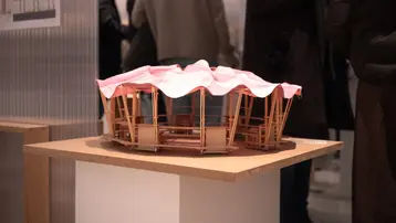 The picture shows an architectural model of an event venue with a round, protective roof made of pink and white striped fabric. It stands on a light-colored table in an exhibition, surrounded by people chatting.