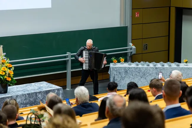 Ein Mann spielt Akkordeon auf einer Bühne in einem Vortragssaal. Im Hintergrund stehen Blumenarrangements und eine lange Tischreihe. Das Publikum sitzt auf Holzstühlen und schaut aufmerksam in seine Richtung.