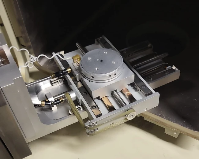 Das Bild zeigt eine präzise Maschinenkomponente mit einem runden Tisch und mehreren beweglichen Teilen. Die Struktur besteht aus Metall und ist für technische Anwendungen konzipiert. Ein Kabel ist sichtbar, das mit der Anlage verbunden ist.