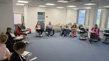 Eine Gruppe von Menschen sitzt in einem modern ausgestatteten Raum und diskutiert. Tische sind in einem halbkreisförmigen Arrangement angeordnet, während einige Personen Notizen machen oder auf ihre Papiere schauen. Im Hintergrund sind Snacks und Getränke auf einem Tisch vorbereitet.