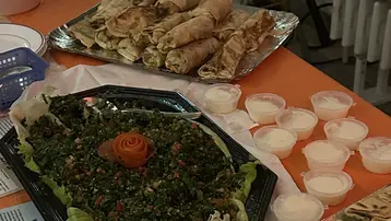 Ein Tisch mit verschiedenen Speisen: eine große Platte mit Taboulé, dekoriert mit einer Karotte in der Mitte, und zwei Sorten gefüllte Teigröllchen. Daneben befinden sich kleine Schalen mit Desserts und ein Stapel Teller. Informative Broschüren liegen ebenfalls auf dem Tisch.