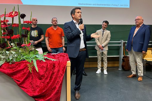 Ein Mann mit Mikrophon spricht zum Publikum. Dahinter stehen weitere Personen.